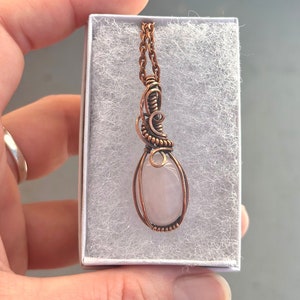 May include: A delicate rose quartz pendant wrapped in copper wire, hanging from a copper chain. The pendant is in a white box with a soft, white lining.