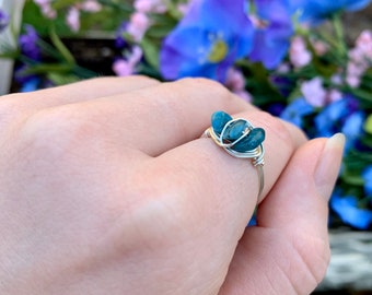May include: A silver wire wrapped ring with two blue-green stones. The ring is on a hand with a blurred background of flowers.