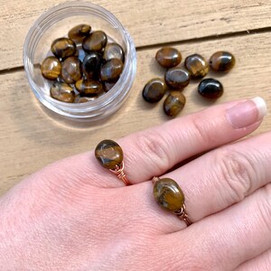 May include: Two rings made with copper wire and tiger's eye stones. The rings are on a person's finger. There are also loose tiger's eye stones in a clear container.