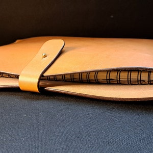 May include: A light brown leather notebook cover with a spiral-bound notebook inside. The cover has a strap closure with a gold-colored button. The notebook is visible inside the open cover.