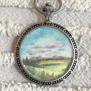 May include: A silver necklace with a round pendant featuring a watercolor painting of a landscape with a blue sky, pink clouds, and green hills.