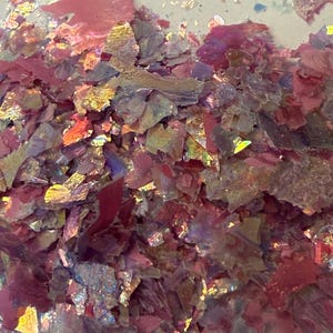 May include: Close-up of iridescent, multi-colored flakes. The flakes are primarily shades of pink, red, and gold, with a shimmering, reflective quality. The texture appears irregular and fragmented, creating a dynamic visual effect.