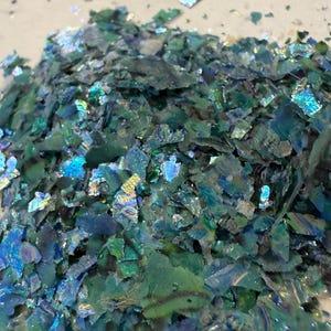 May include: A pile of iridescent, crushed flakes in shades of teal, blue, and green. The flakes have a reflective, shimmering quality, catching the light to create a sparkling effect. The material appears to be a decorative craft supply.