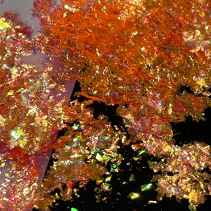 May include: Orange and gold iridescent glitter flakes on a black background. The glitter is scattered and has a variety of sizes and shapes.