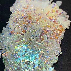 May include: A close-up of a piece of iridescent glass with a white background. The glass has a variety of colors, including blue, green, orange, and red. The glass is broken into small pieces, creating a mosaic effect.