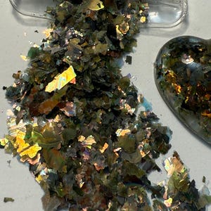 May include: Iridescent green and gold flakes spilling from a clear container. The flakes have a shimmering, reflective quality, with hints of yellow and purple. A heart-shaped object is visible in the background, also filled with the same flakes.
