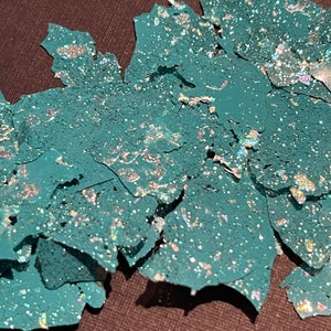 May include: Teal glitter flakes with iridescent shimmer. The flakes are scattered on a dark surface.