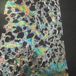 May include: A rectangular, iridescent sheet with a network of irregular holes. The surface reflects a spectrum of colors, including blues, greens, yellows, and purples. The material appears translucent, with a textured, porous structure.