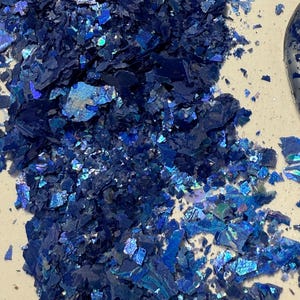 May include: A pile of iridescent blue flakes with a reflective, textured surface. The flakes vary in size and shape, creating a shimmering effect. The color shifts from deep navy to lighter shades of blue.