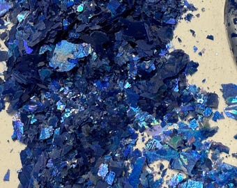 Puede incluir: Un montón de escamas azules iridiscentes con una superficie reflectante y texturizada. Las escamas varían en tamaño y forma, creando un efecto brillante. El color cambia de azul marino oscuro a tonos de azul más claros.