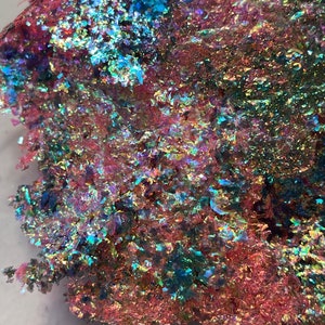 May include: A close-up of iridescent glitter flakes in shades of pink, blue, and green. The glitter is scattered on a white surface.