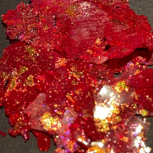 May include: Red and gold glitter flakes, scattered on a black surface. The glitter is iridescent and has a variety of sizes and shapes.