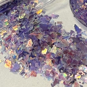 May include: Close-up of iridescent purple and multicolored flakes scattered on a light surface. The flakes have a reflective quality, with hints of gold, pink, and green. The flakes are in a clear container.