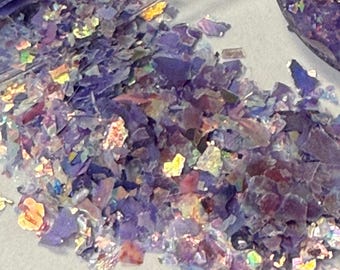 Puede incluir: Primer plano de copos iridiscentes morados y multicolores esparcidos sobre una superficie clara. Los copos tienen una cualidad reflectante, con toques de dorado, rosa y verde. Los copos están en un recipiente transparente.