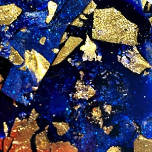 May include: Close-up of a blue and gold resin art piece with a glittery finish. The resin is layered with gold flakes and has a textured surface.