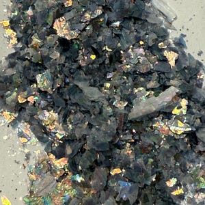 May include: Close-up of a pile of dark, iridescent flakes. The flakes have a layered appearance with a mix of dark gray and shimmering, colorful highlights. The overall texture appears rough and fragmented.