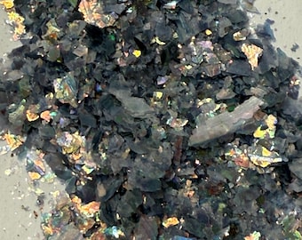 May include: Close-up of a pile of dark, iridescent flakes. The flakes have a layered appearance with a mix of dark gray and shimmering, colorful highlights. The overall texture appears rough and fragmented.