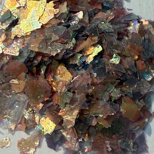 May include: Close-up of iridescent flakes in shades of gold, bronze, and green. The flakes have a metallic sheen and a textured, irregular shape. The material appears to be scattered on a light-colored surface.