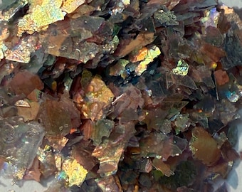 Puede incluir: Primer plano de escamas iridiscentes en tonos dorados, bronce y verde. Las escamas tienen un brillo metálico y una forma irregular texturizada. El material parece estar disperso sobre una superficie de color claro.