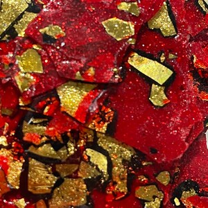 May include: Close-up of a red and gold abstract art piece. The piece is made of resin and has a textured surface. The gold is in the shape of flakes and is scattered throughout the piece.