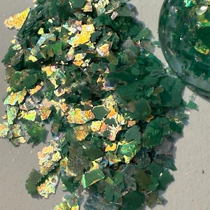 May include: Close-up of green and iridescent glitter flakes. The flakes have a textured, irregular shape and reflect multiple colors. A clear, round object filled with similar glitter is also visible.