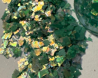 Puede incluir: Primer plano de escamas de purpurina verdes e iridiscentes. Las escamas tienen una forma irregular y texturizada y reflejan múltiples colores. También se ve un objeto redondo y transparente lleno de purpurina similar.