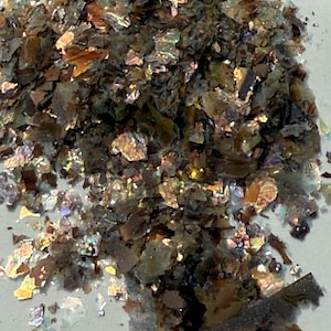 May include: Close-up of a pile of iridescent flakes. The flakes are primarily brown and gray, with shimmering, colorful highlights of gold, green, and purple. The texture appears rough and fragmented, suggesting a natural material.