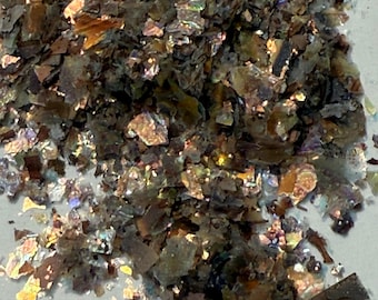 May include: Close-up of a pile of iridescent flakes. The flakes are primarily brown and gray, with shimmering, colorful highlights of gold, green, and purple. The texture appears rough and fragmented, suggesting a natural material.