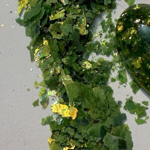 May include: Close-up of green and gold iridescent flakes. The flakes are scattered across a light gray surface. The flakes have a layered appearance and reflect light, creating a shimmering effect. A rounded object with a similar color scheme is visible in the background.