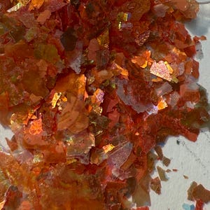 May include: Close-up of iridescent orange and red flakes. The flakes have a shattered appearance, with sharp edges and varying sizes. The colors shift with the light, creating a shimmering effect. The flakes appear to be a decorative material.