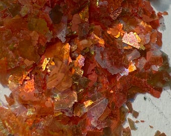 Puede incluir: Primer plano de escamas iridiscentes de color naranja y rojo. Las escamas tienen una apariencia fragmentada, con bordes afilados y tamaños variables. Los colores cambian con la luz, creando un efecto brillante. Las escamas parecen ser un material decorativo.