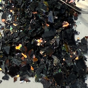 May include: Close-up of a pile of black, iridescent flakes. The flakes have a layered appearance and reflect a spectrum of colors, including gold, green, and blue. The material appears to be a decorative element, possibly for crafts or nail art.