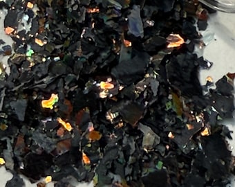 May include: Close-up of a pile of black, iridescent flakes. The flakes have a layered appearance and reflect a spectrum of colors, including gold, green, and blue. The material appears to be a decorative element, possibly for crafts or nail art.