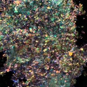 May include: A close-up of iridescent green, blue, and gold glitter. The glitter is scattered on a black surface.