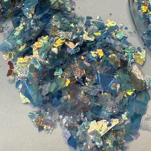 May include: A pile of iridescent blue and gold flakes. The flakes are irregularly shaped and reflect light, creating a shimmering effect. The flakes appear to be scattered on a white surface.