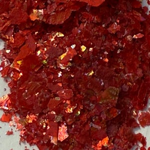 May include: Close-up of a pile of red, irregularly shaped flakes. The flakes have a glossy, reflective surface with hints of gold and orange. The overall texture appears rough and fragmented, with varying sizes and shapes.