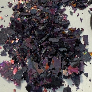 May include: A pile of dark purple, iridescent flakes with hints of red, orange, and silver. The flakes have a textured, irregular shape and are scattered across a light gray surface. The material appears to be decorative.