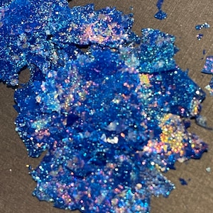 May include: A pile of iridescent blue glitter with a holographic finish. The glitter is scattered on a dark gray surface.