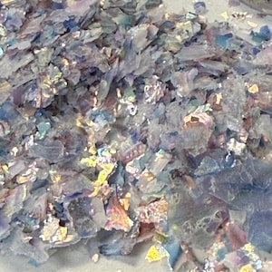 May include: Close-up of iridescent, multi-colored flakes. The flakes have a shimmering, reflective quality, with hues of blue, purple, and gold. The material appears to be thin and fragmented, possibly for crafting or decorative purposes.