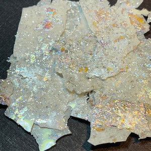 May include: A pile of iridescent glitter flakes on a black surface. The glitter is white with a rainbow sheen.