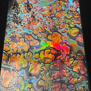 May include: A rectangular sheet with a vibrant, iridescent pattern. The design features a mix of colors, including orange, yellow, green, and blue, with a glossy, reflective surface. The abstract shapes create a dynamic and eye-catching visual effect.