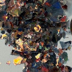 May include: A pile of iridescent, multi-colored flakes. The flakes have a metallic sheen, with colors ranging from deep blues and greens to fiery oranges and reds. The fragments are irregularly shaped and appear to be scattered across a light-colored surface.