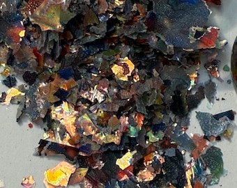 May include: A pile of iridescent, multi-colored flakes. The flakes have a metallic sheen, with colors ranging from deep blues and greens to fiery oranges and reds. The fragments are irregularly shaped and appear to be scattered across a light-colored surface.