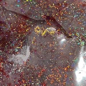 May include: Close-up of a red, white, and gold glitter resin art piece. The glitter is iridescent and creates a shimmering effect. The resin is clear and allows the glitter to shine through.