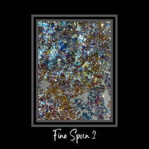 May include: Close-up of iridescent, fine-spun glitter with a blue, green, and gold color scheme. The glitter is arranged in a random pattern and appears to be scattered on a white surface. The text "Fine Spun 2" is written below the image.