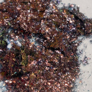 May include: A close-up of iridescent copper flakes, with a variety of sizes and shapes. The flakes have a metallic sheen and reflect light in a rainbow of colors.