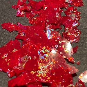 May include: Red iridescent glitter flakes, scattered on a dark gray surface. The glitter has a holographic finish, with shades of pink, blue, and green.