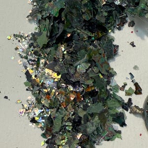 May include: A pile of iridescent green and black flakes. The flakes have a metallic sheen and reflect multiple colors, including gold, silver, and blue. The flakes are scattered on a white surface.