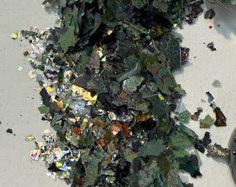 May include: A pile of iridescent green and black flakes. The flakes have a metallic sheen and reflect multiple colors, including gold, silver, and blue. The flakes are scattered on a white surface.