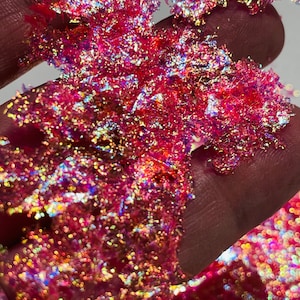 May include: Close-up of iridescent pink glitter flakes. The glitter has a reflective, shimmering quality, with hints of gold, blue, and purple. The flakes are clustered together, creating a textured appearance. The glitter is likely used for crafts or decorative purposes.